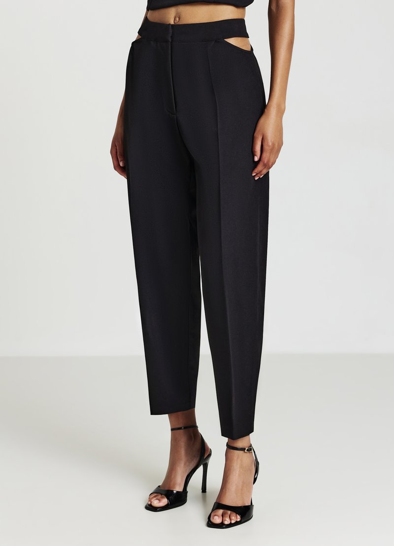 Pantalons noirs taille haute avec découpes latérales, coupe ajustée, texture lisse et jambes fuselées. Associés à des sandales noires à talons avec lanières.