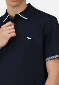 Polo navy in tessuto morbido, con un logo di bassotto bianco sul petto e dettagli a righe sul colletto e le maniche.