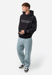 Hanorac negru cu text brodat, blugi deschiși la culoare cu croială lejeră și adidași alb-negru. Gluga căptușită; eticheta brandului vizibilă pe manșetă.