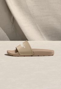 Μπεζ σανδάλι τύπου slide με φαρδύ λουρί που φέρει λευκό κείμενο, επίπεδη σόλα και ελαφρώς υφασμένη επιφάνεια, φωτογραφημένο σε φωτεινό ουδέτερο φόντο.