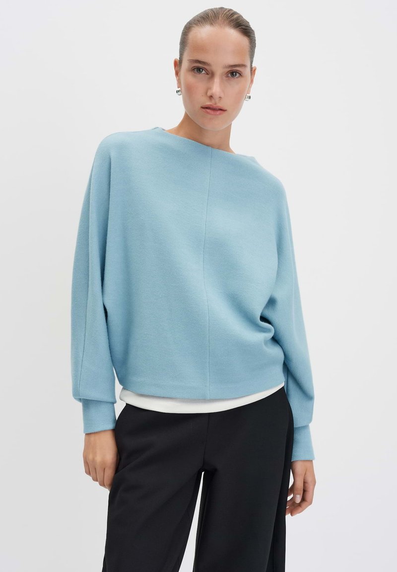 Un pull surdimensionné bleu clair en matériau doux, avec un col bateau et des épaules tombantes, associé à un pantalon noir à jambes larges.