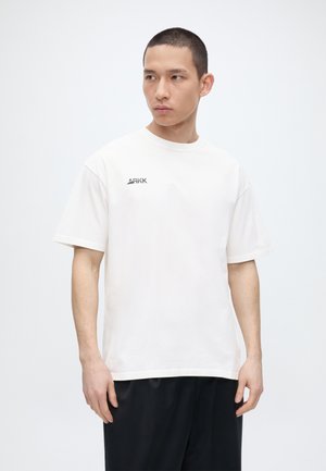 Junger Mann mit kurzen Haaren, trägt ein weißes ARKK-T-Shirt und schwarze Hose, schaut nach links vor einem einfarbig hellen Hintergrund.