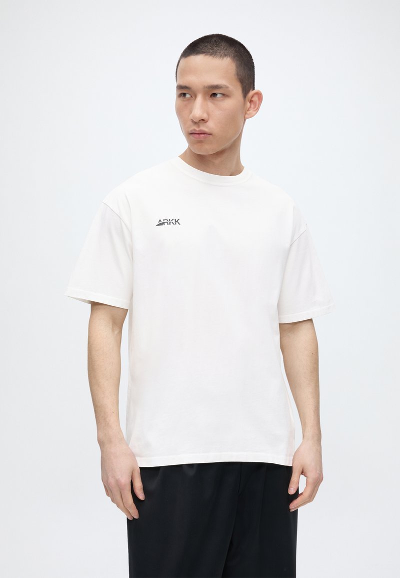 Νεαρός άνδρας με κοντά μαλλιά που φοράει λευκό t-shirt ARKK και μαύρο παντελόνι, κοιτάζοντας αριστερά σε απλό ανοιχτό φόντο.