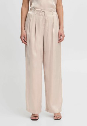 Lichtbeige broek met wijde pijpen van een gladde, satijnachtige stof; met plooien en een strikceintuur. Gepaarde met zwarte, bandjes sandalen.