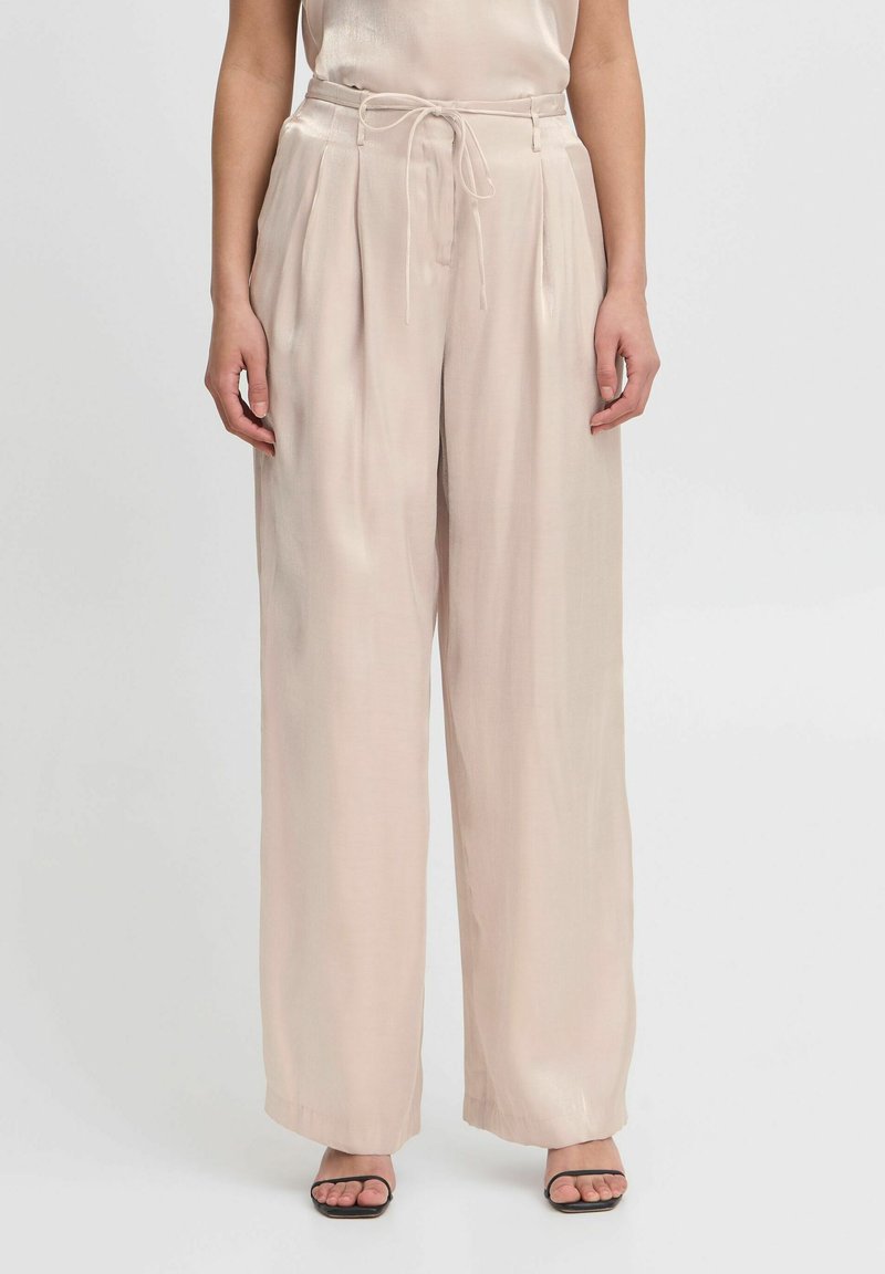 Lichtbeige broek met wijde pijpen van een gladde, satijnachtige stof; met plooien en een strikceintuur. Gepaarde met zwarte, bandjes sandalen.