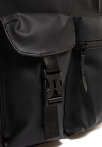 Mochila negra fabricada con material impermeable, con tapa de cierre, correa ajustable y bolsillo con cremallera con detalles texturizados.