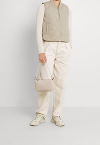 Beżowa pikowana kamizelka na kremowej prążkowanej bluzce, w połączeniu z jasnobeżowymi spodniami i dopasowaną małą torebką. Uzupełnia to kremowe trampki.