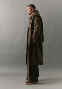 Hosszú barna és zöld hounds-tooth kabát, oversized barna sálat viselve; egy barna kapucnis felsőre és sötét nadrágra rétegezve. Vastag barna lábbeli.