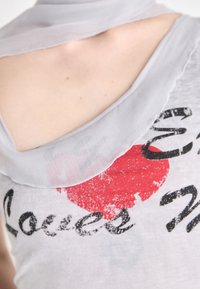 Top sans manches gris avec un col arrondi, arborant un graphique de cœur rouge et un texte noir indiquant "Loves Me". Tissu doux et léger.