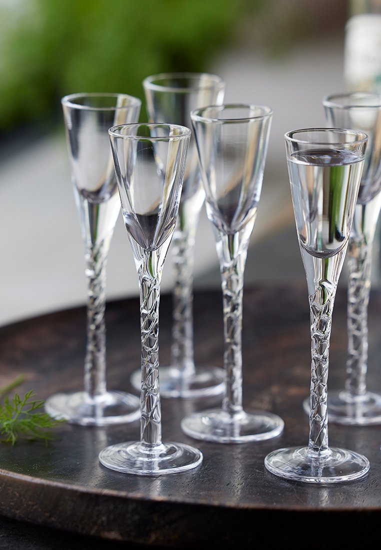 Przezroczyste szklane kieliszki do shotów z wysokimi, teksturowanymi nóżkami i szerokimi podstawami, ułożone na ciemnej, drewnianej tacy. Kieliszki zawierają klarowny płyn.