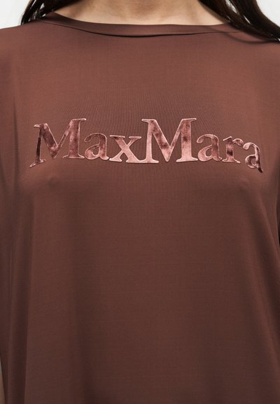 Chemise en tissu marron avec le logo "MaxMara" en velours rose texturé sur la poitrine, portée par une personne aux longs cheveux foncés.