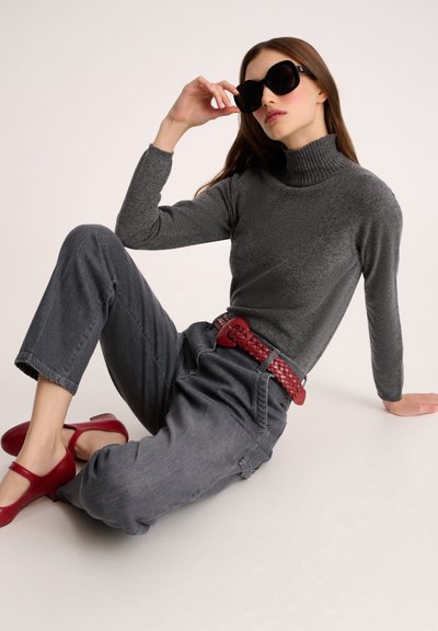 Grauer Rollkragenpullover, graue weit geschnittene Jeans, geflochtener roter Gürtel und rote Schuhe. Modell trägt große schwarze Sonnenbrille und sitzt mit angezogenem Bein.