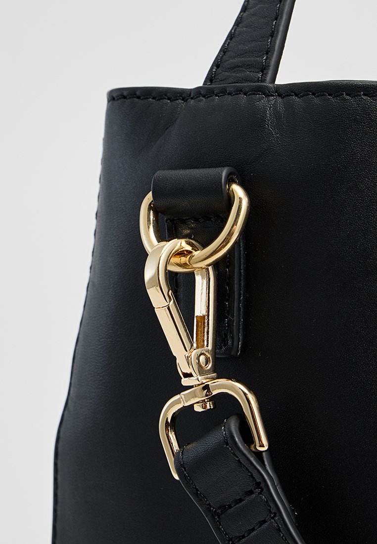 Angolo di una borsa in pelle nera con una chiusura in metallo dorato e un anello, caratterizzata da dettagli cuciti nella tracolla. Texture liscia con una sottile lucentezza.