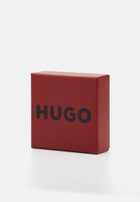 Červená štvorcová krabička vyrobená z textúrovaného papiera. Na prednej strane sú tučné čierne písmená, ktoré tvoria slovo "HUGO". Jednoduchý dizajn s hladkým povrchom.