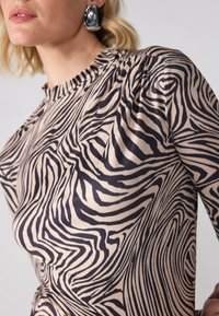 T-shirt à manches longues avec une base beige ornée de rayures noires de zèbre. Fabriqué en tissu lisse avec uncol en gathered et une texture subtile.