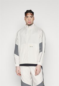 White zip-up jacket with gray accents, featuring a faint logo. The fabric appears lightweight with elastic cuffs and a relaxed fit.