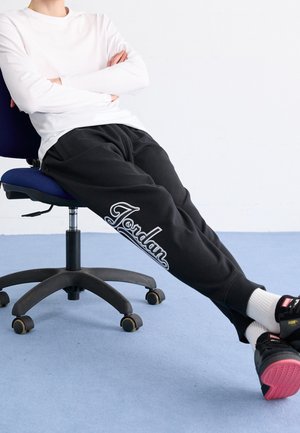 Personne portant un pantalon de jogging Jordan noir, une chemise blanche à manches longues et des baskets noires assise sur une chaise de bureau bleue sur un sol bleu.
