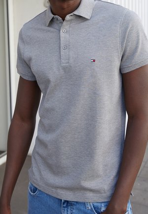 Polo gris avec col, trois boutons et un petit logo aux couleurs rouge, blanc et bleu. Tissu doux ; manches courtes ; coupe décontractée.