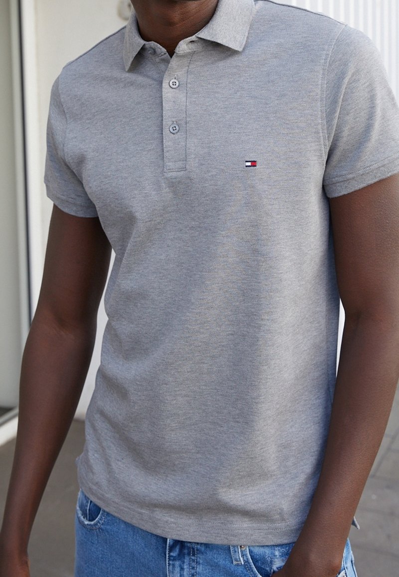 Polo gris avec col, trois boutons et un petit logo aux couleurs rouge, blanc et bleu. Tissu doux ; manches courtes ; coupe décontractée.
