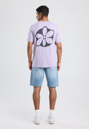 T-shirt lilas à manches courtes avec un grand motif floral noir au dos. Associé à un short en denim bleu clair et des baskets blanches.