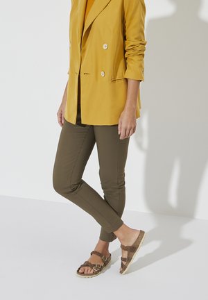 Persoon in olijfgroene broek, een mosterdgele blazer met opgerolde mouwen en bruine kurken sandalen met dubbele banden, staande tegen een witte achtergrond.