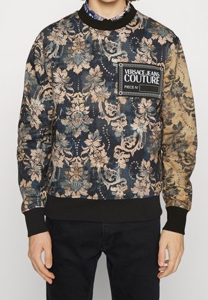 Personne portant un sweat-shirt Versace Jeans Couture à motif floral avec des poignets et un ourlet noirs, associé à un pantalon noir.