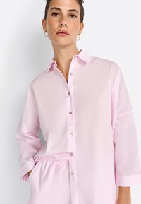 Light pink shirt with a button-up front, long sleeves, and a relaxed fit, featuring shiny buttons and a rounded collar.