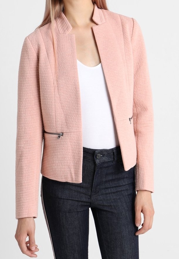 Blazer texturé rose pâle avec un col montant et une ouverture avant. Comprend deux poches zippées et une coupe moderne et cintrée. Porté sur un haut blanc.