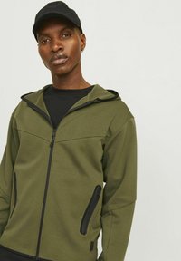 Haut à capuche zippé vert olive avec accents noirs ; comporte une fermeture éclair à l'avant, deux poches latérales zippées et une capuche. Tissu léger.
