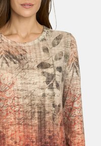 Top de manga larga con estampado floral y escote redondeado. Los colores se mezclan desde el beige hasta el óxido, presentando diseños de escritura y hojas sobre un tejido texturizado.