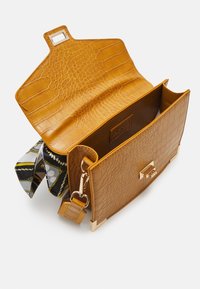 Sac bandoulière en cuir crocodile marron clair avec ferrures dorées, rabat ouvert et tissu à motifs noir, blanc et jaune visible à l'intérieur.