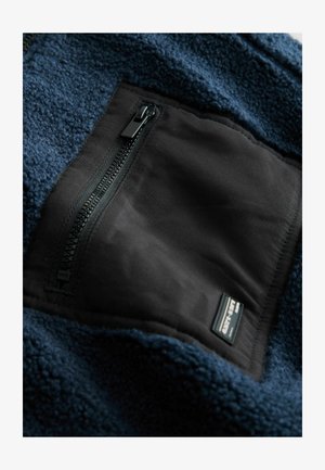 Tumes sinine fliistekstiil, millel on tekstuuriga viimistlus, musta tõmblukuga tasku ja ristkülikukujuline silt kangal.