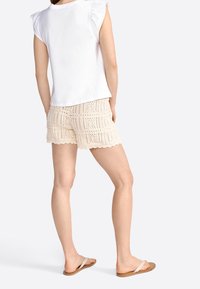 Top de algodón blanco con hombros con volantes, combinado con pantalones cortos de croché beige que presentan un dobladillo con ondas y patrones cuadrados texturizados.