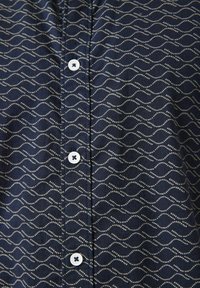 Chemise bleu marine avec un motif de vagues en points beiges, conceptrice à boutons avec des boutons blancs, tissu texturé pour plus de détail.