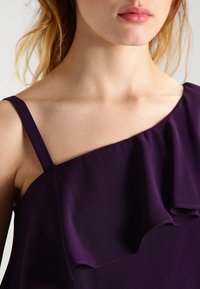 Un primo piano di un indumento viola a spalla scoperta con un design volante e spalline sottili, realizzato in un tessuto liscio e morbido.