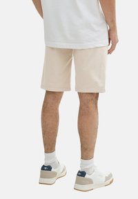 Beige Baumwollshorts mit lockerer Passform und gesäumtem Saum, kombiniert mit weißen Sneakers und weißen Socken. Einfaches, sauberes Design.