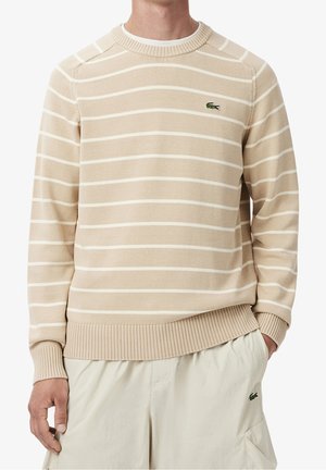 Beiger Pullover mit Rundhalsausschnitt und weißen horizontalen Streifen, getragen von einer Person mit der Hand in der Tasche, kombiniert mit hellbeigen Hosen.