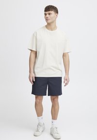 Krém színű pamut póló rövid ujjakkal és kerek nyakkivágással, sötétkék rövidnadrággal és fehér sportcipővel párosítva, egyszerű háttér előtt állva.