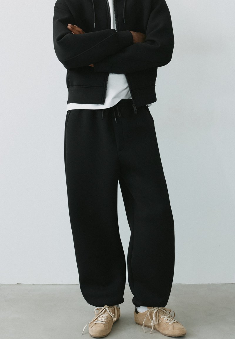 Personne portant un sweat à capuche noir zippé, un pantalon de survêtement noir, un maillot blanc et des baskets beiges, debout les bras croisés.