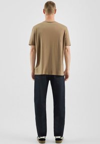 T-shirt marron à col rond associé à un jean bleu foncé droit et des baskets noires et blanches, vu de dos. Texture en coton lisse.