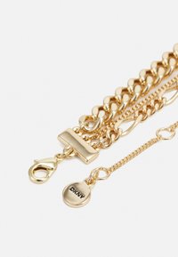 Bracciale dorato in tonalità oro con maglie a catena intrecciate, chiusura a moschettone e un'etichetta circolare con inciso "DKNY". Finitura liscia e lucida.