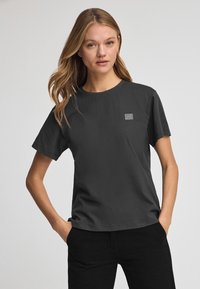 Mörkgrå bomullströja med rund halsringning, korta ärmar och en liten rektangulär logga på bröstet. Slät textur, avslappnad passform.