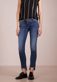 Pantalones vaqueros oscuros y ajustados con un ligero desgaste, dobladillo deshilachado y detalles plateados en los laterales. Combinados con una blusa negra de manga corta con detalles dorados.