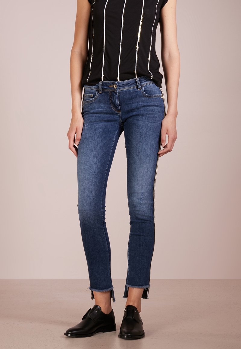 Pantalones vaqueros oscuros y ajustados con un ligero desgaste, dobladillo deshilachado y detalles plateados en los laterales. Combinados con una blusa negra de manga corta con detalles dorados.