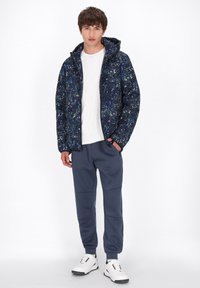 Mariniersjack met een meerkleurig spetterpatroon, ritsluiting en capuchon; gecombineerd met grijze joggers en witte sneakers.