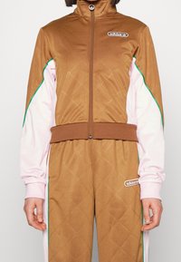 Brown and pink Adidas tracksuit with green piping and logo patches on chest and thigh, worn by a standing person.