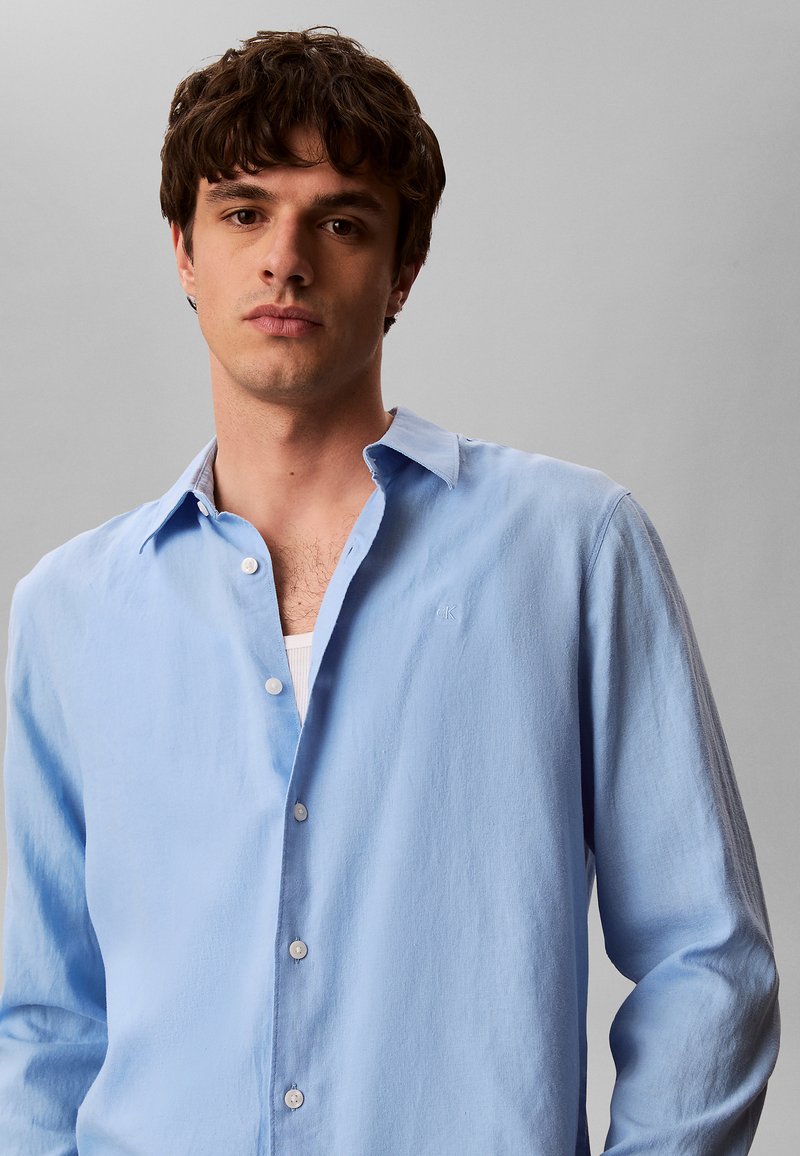 Jonge man met kort bruin haar, gekleed in een lichtblauw overhemd met knopen over een wit ondershirt, staand voor een effen grijze achtergrond.