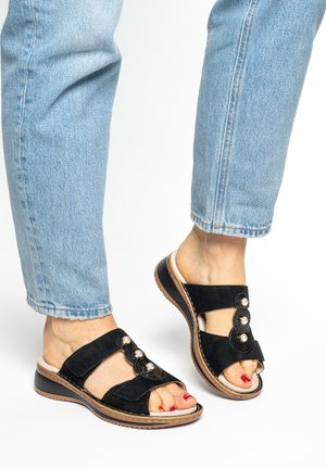 Black suede sandals with an open toe, featuring a decorative design of three beads and a textured sole. Worn with light blue jeans.