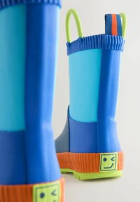 Blaue und hellblaue Gummiregenstiefel mit einer strukturierten orangen Sohle, grünen Akzenten und einem Smiley-Gesicht-Detail an der Ferse.