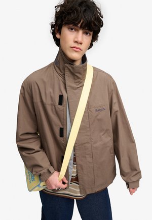 Ung person med krøllet mørkt hår iført en brun Bench jakke, stribet skjorte, blå bukser og bærende på en beige crossbody taske.